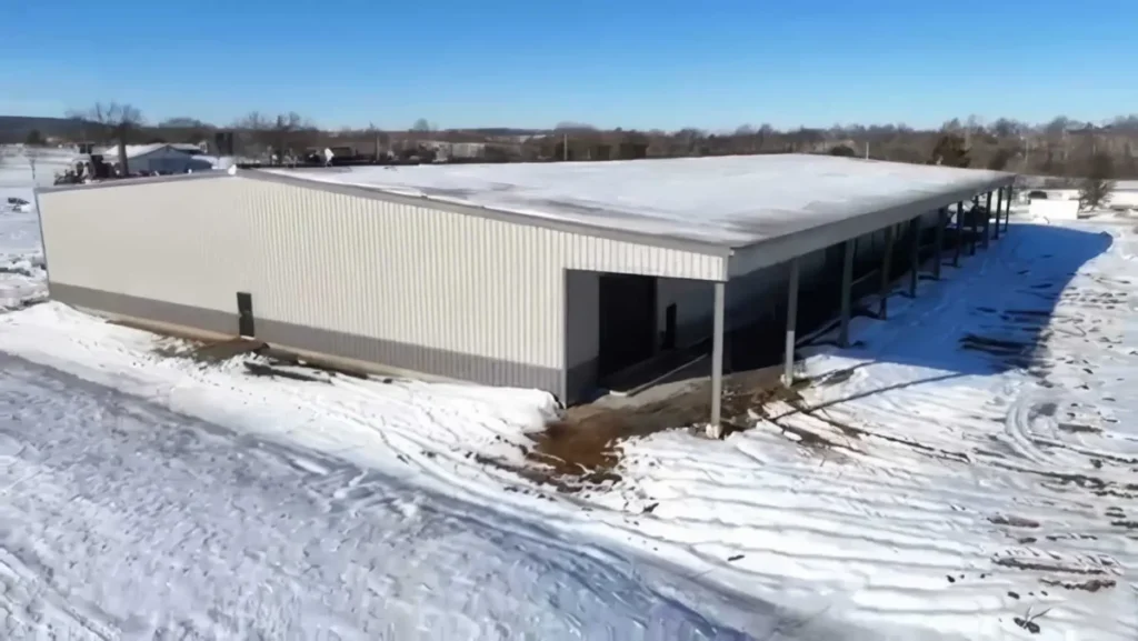 Aerial drone view of a large steel metal building with a tan corrugated exterior and snow-covered flat roof, surrounded by a snowy rural landscape under a clear blue sky.