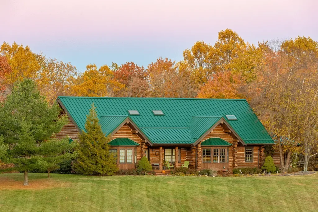 A large log cabin with a green standing-seam metal roof and skylights sits in front of vibrant autumn foliage, surrounded by a manicured green lawn and evergreen trees.