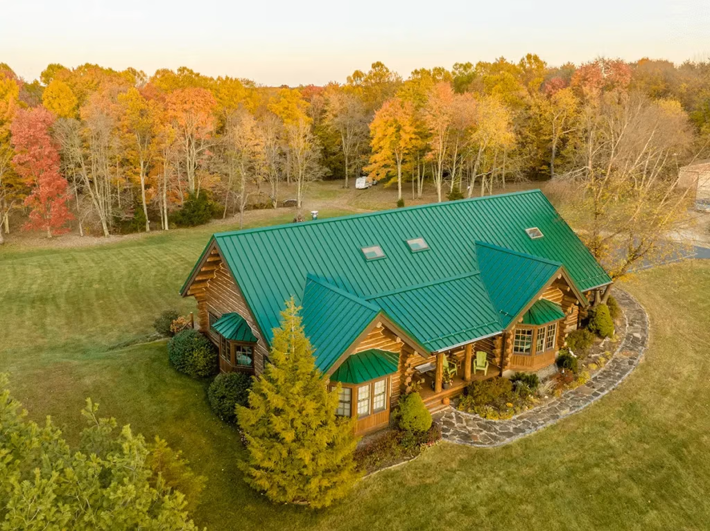 Aerial view of a large log cabin with a bright green standing-seam metal roof surrounded by manicured landscaping and vibrant fall foliage in red, orange, and yellow.