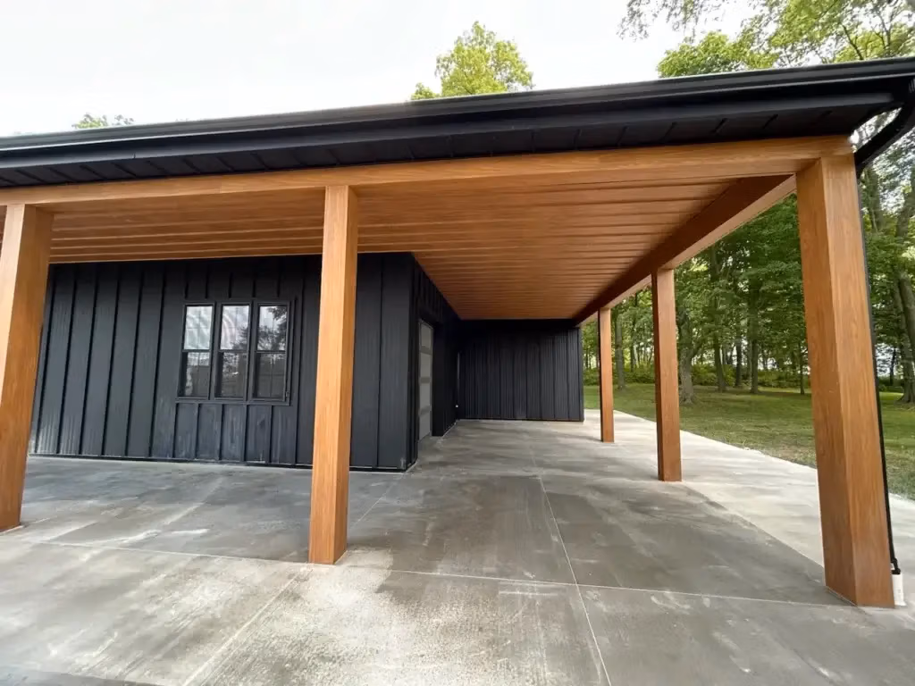 A modern black vertical steel panel barndominium featuring a covered carport area with warm wood-grain painted posts and ceiling, concrete flooring, and a decorative window, surrounded by green trees.