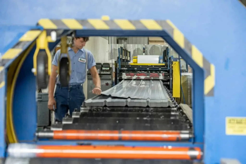 A male factory worker in a blue uniform shirt guides freshly roll-formed corrugated metal roofing panels along a conveyor line inside a blue industrial manufacturing machine.