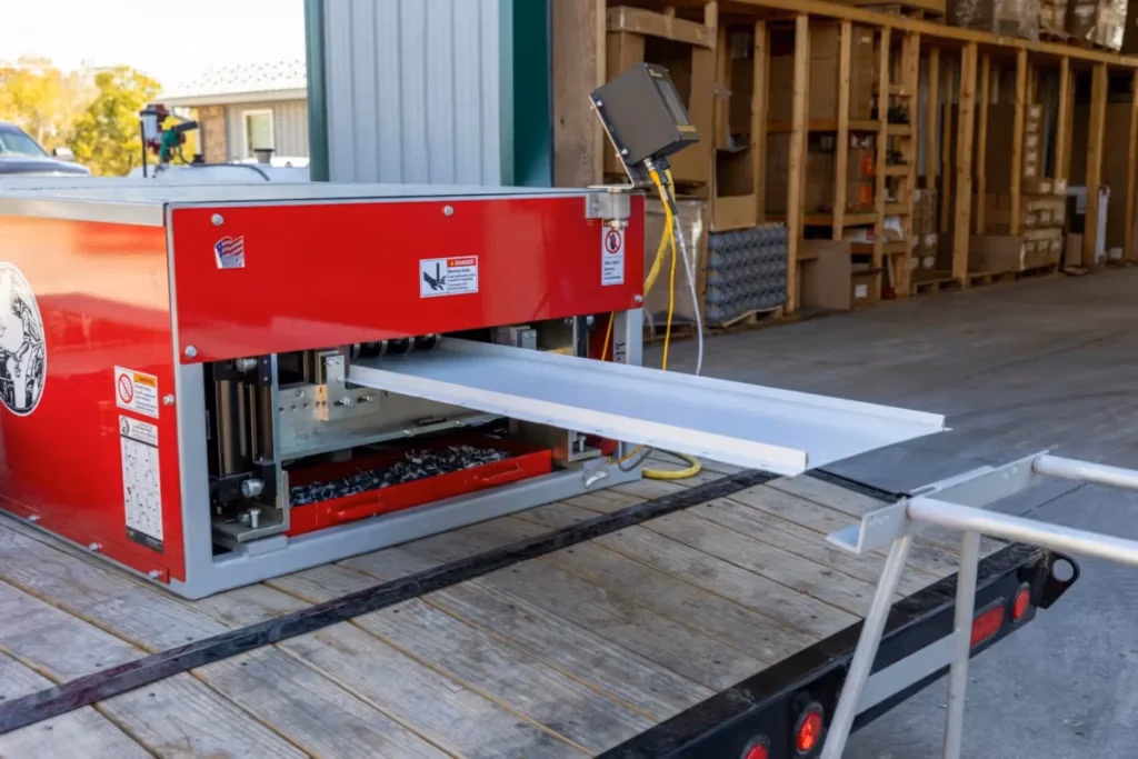 A red portable roll forming machine mounted on a flatbed trailer produces a white metal board and batten trim panel outside an open warehouse building.