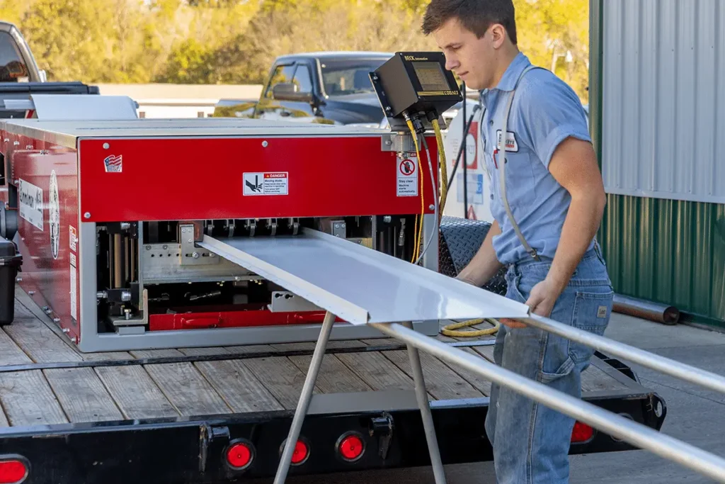 A young male technician in a blue polo shirt guides a white metal panel being produced by a red mobile roll forming machine mounted on a flatbed trailer outside a metal building.