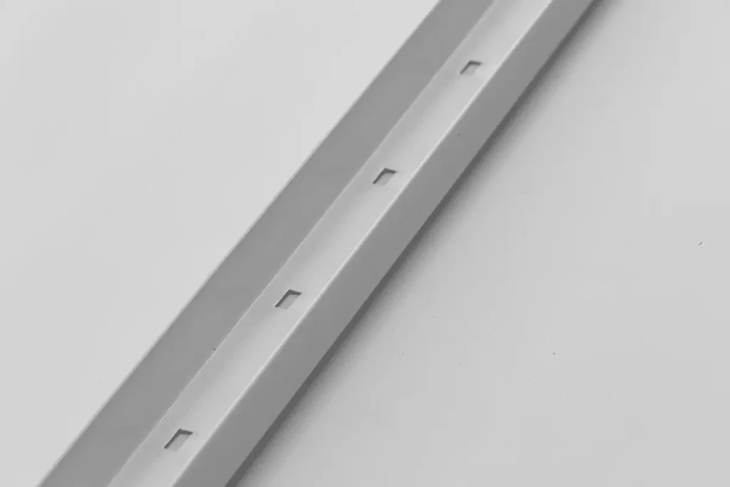 A close-up of a silver aluminum slotted batten strip lying diagonally against a white background, featuring evenly spaced rectangular notches along its center channel.