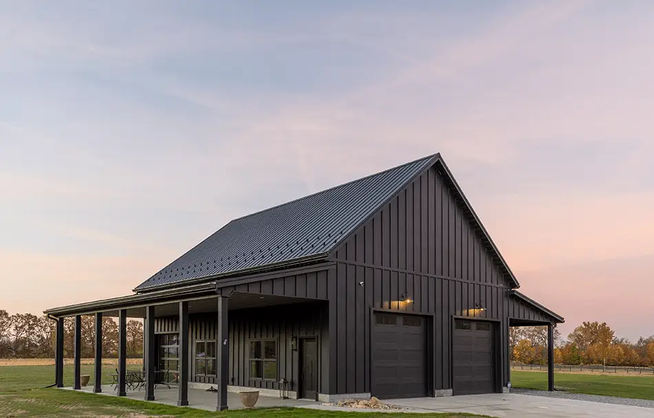 A modern dark charcoal gray barndominium with vertical metal board and batten siding, a standing seam metal roof, covered wraparound porch, and two black garage doors, photographed at dusk on a rural property with green grass and trees in the background.