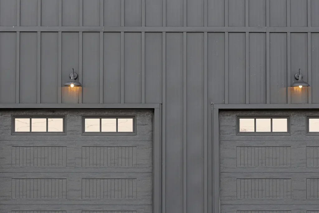 A symmetrical dark charcoal gray metal board and batten exterior wall features two carriage-style garage doors with rectangular transom windows and matching gooseneck barn lights mounted above each door.