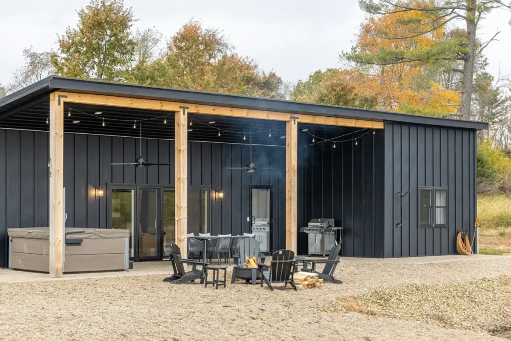 A modern black metal board and batten barndominium with natural wood beam porch posts, string lights, Adirondack chairs around a fire pit, a hot tub, and gas grills, surrounded by autumn trees.