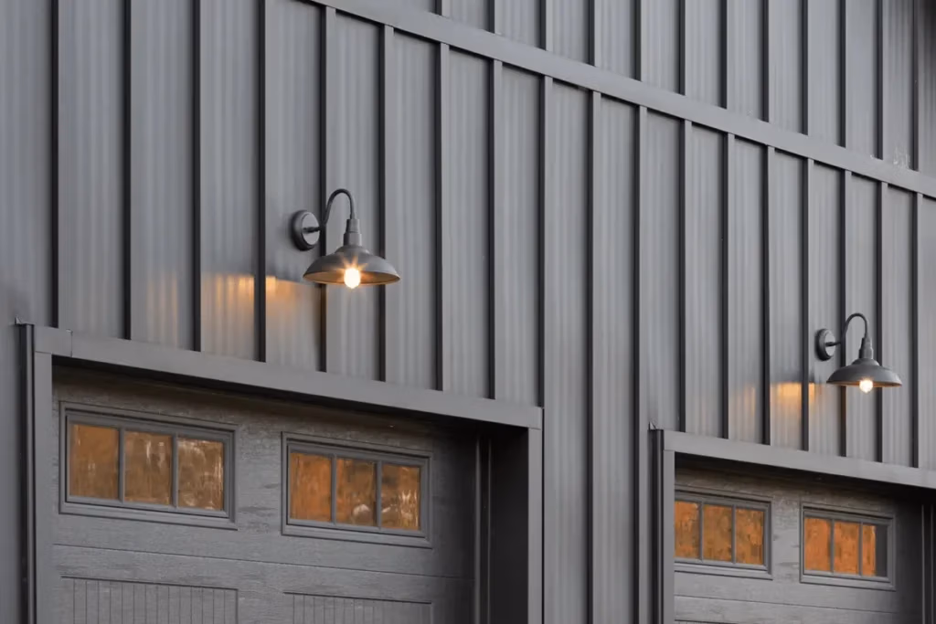 Two dark gray metal board and batten garage doors with glass panel windows are mounted on a corrugated metal wall, each flanked by illuminated black gooseneck barn lights casting a warm amber glow.