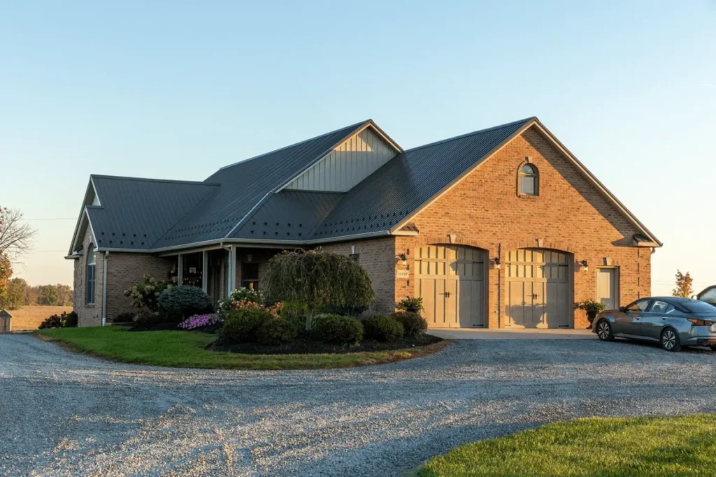 A brick ranch-style home with a dark gray standing seam metal roof, tan carriage-style garage doors, and landscaped front yard, photographed at golden hour with a gravel driveway in the foreground.