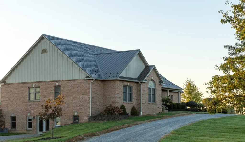 A large two-story brick farmhouse with a gray standing seam metal roof, surrounded by green landscaping and a gravel driveway in a rural setting during early evening light.