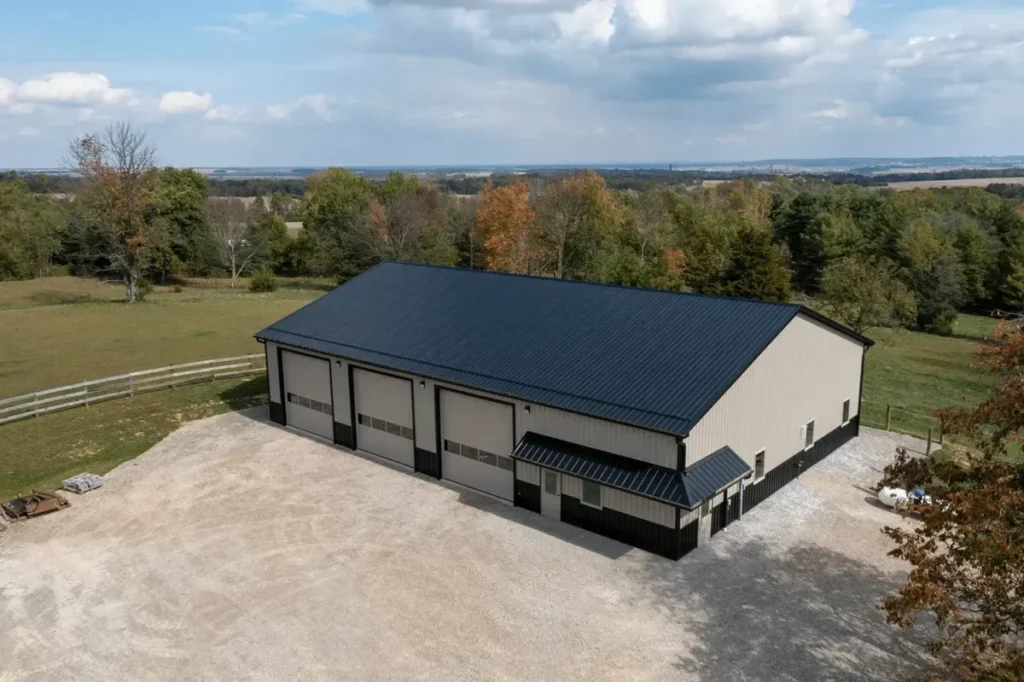 Aerial drone view of a large cream and black metal pole barn with a dark standing-seam roof, three overhead garage doors, and a covered entry awning, surrounded by gravel parking and open farmland in rural Ohio.