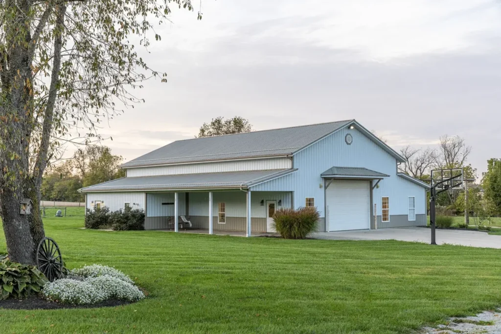 A light blue and gray metal pole barn home with a covered front porch, white garage door, and well-manicured green lawn, flanked by a large tree with a decorative wagon wheel and a basketball hoop on a rural property.