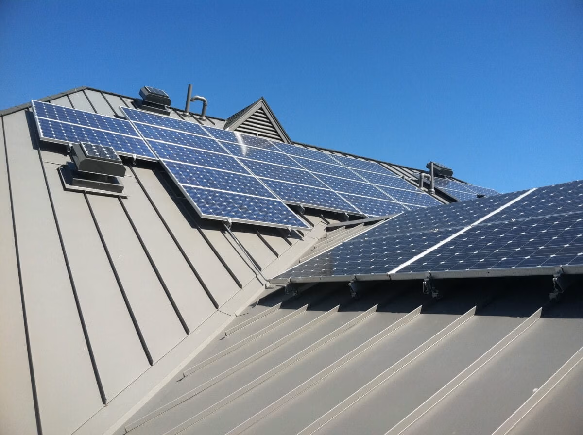 Multiple blue photovoltaic solar panels mounted on a gray standing seam metal roof under a clear blue sky, with roof vents and additional solar arrays visible in the background.