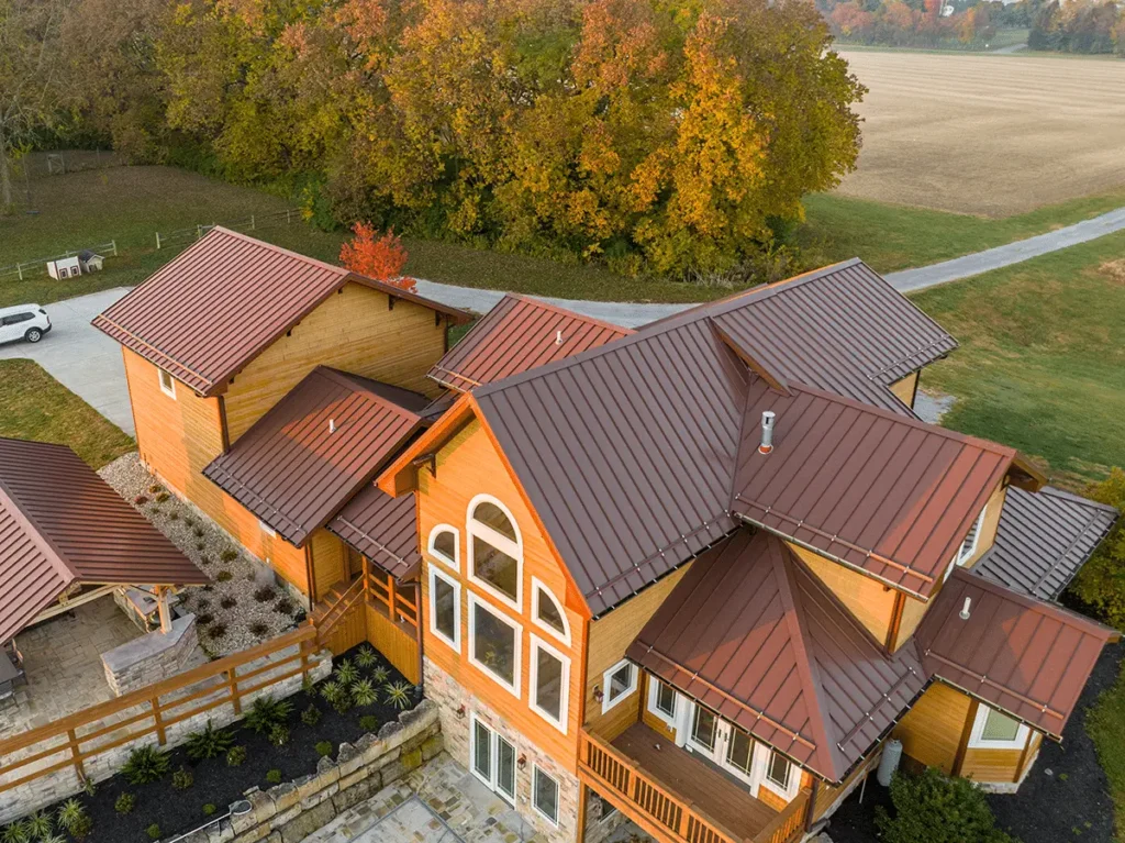 Aerial drone view of a large multi-gabled wood-sided residential home featuring dark reddish-brown standing seam metal roofing, surrounded by autumn foliage and open farmland in rural Ohio.