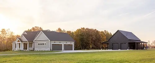 A light gray craftsman-style ranch house with a three-car garage sits beside a dark charcoal metal-roofed barn structure on a green lawn surrounded by autumn trees at golden hour.