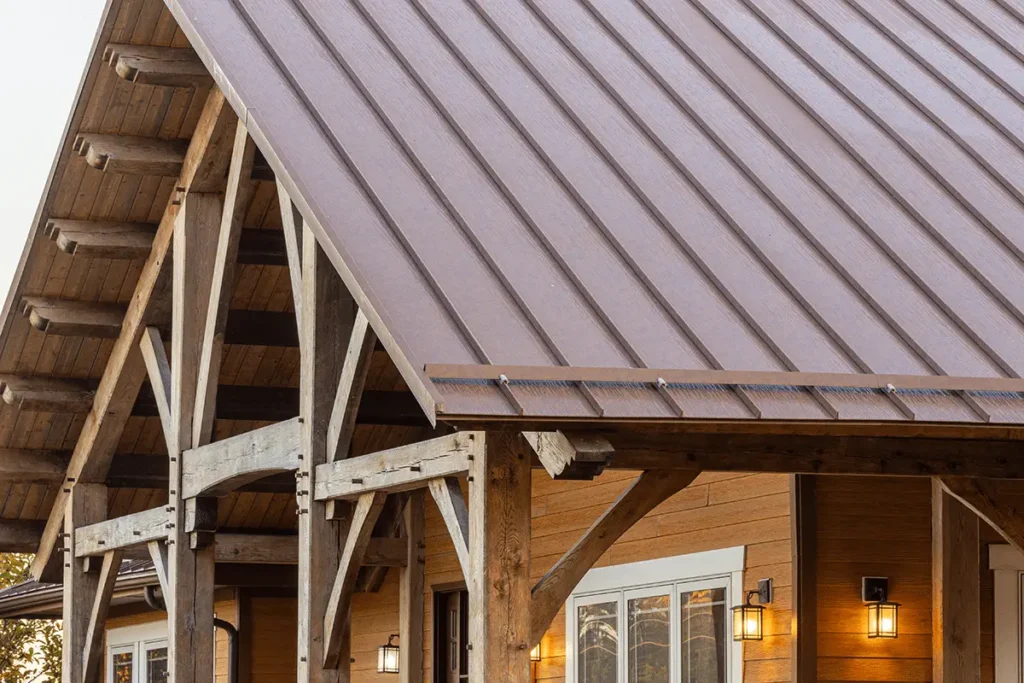 A close-up view of a gray-brown standing seam metal roof crowning a rustic timber frame porch with weathered wood posts, decorative knee braces, and warm lantern-style wall sconces on horizontal wood siding.