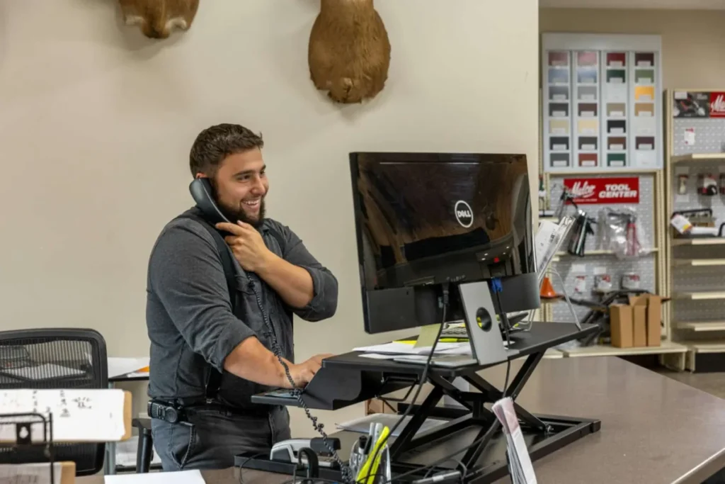 A smiling young man with a beard talks on a corded phone while standing at a sit-stand desk with a Dell monitor in a metal sales showroom office.