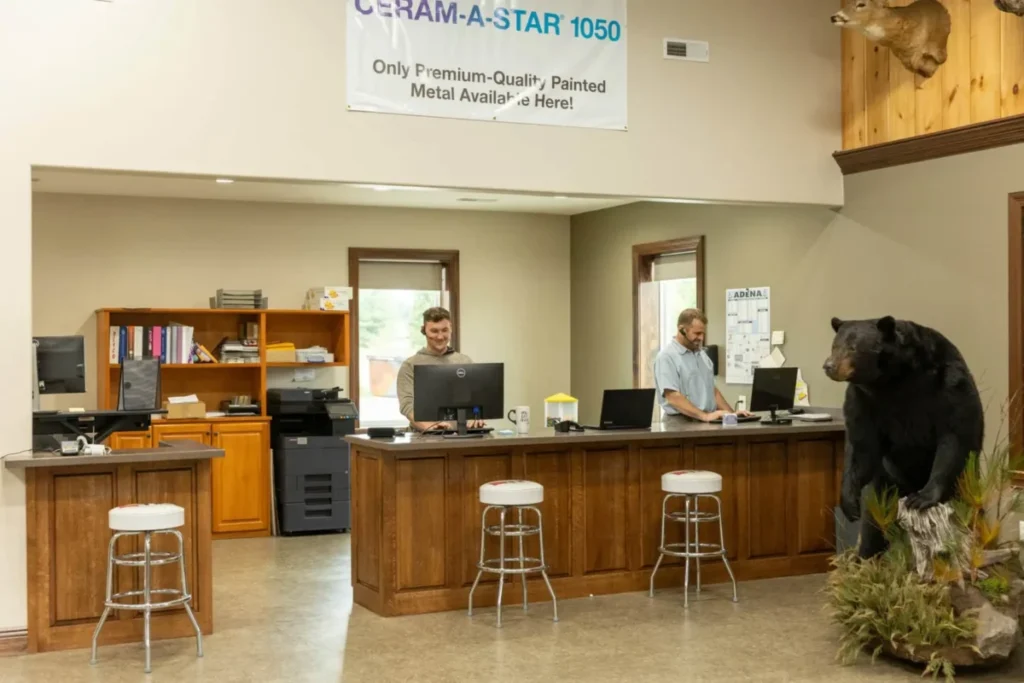 Two male employees work at a wooden customer service counter with computers inside the Buckeye Metal Sales showroom, with a full-body black bear taxidermy mount and a deer head on the wall, beneath a Ceram-A-Star 1050 banner advertising premium-quality painted metal.