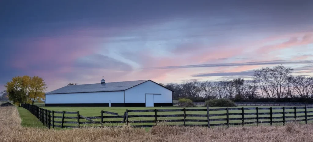 A large white metal pole barn with a dark gray roof and black trim sits behind black wooden paddock fencing on a rural farm property at twilight, with a colorful pink and purple sky overhead and autumn trees in the background.
