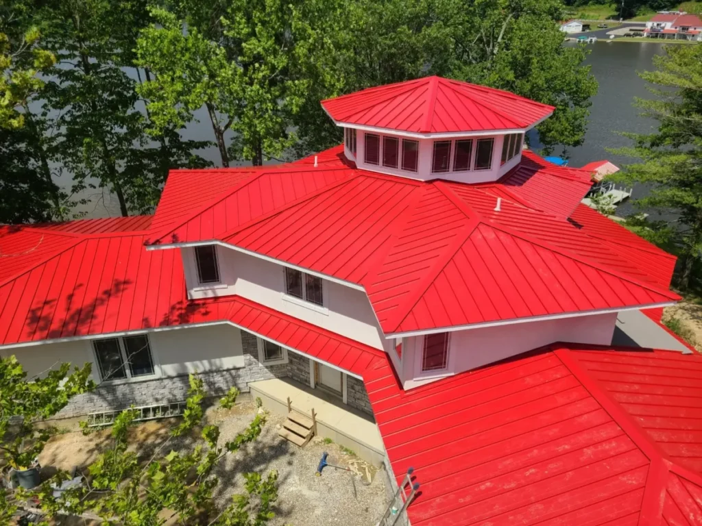 Aerial drone view of a large lakeside house featuring a vibrant red standing seam metal roof with a central octagonal cupola, white fascia, and stone accents, surrounded by green trees near a waterway.