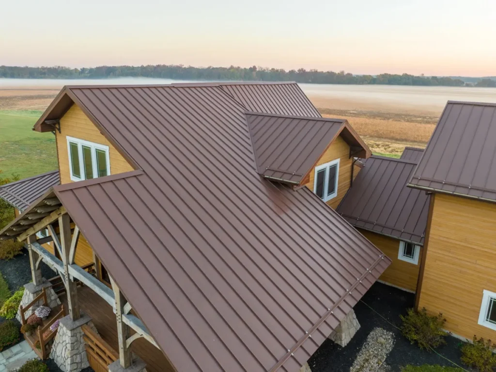 Aerial drone view of a large craftsman-style home with brown standing seam metal roofing, yellow wood siding, and white-framed windows, set against a misty rural Ohio landscape at sunrise.