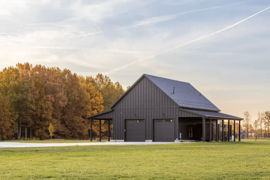 A large black metal board and batten sided barn with two dark garage doors and covered lean-to extensions sits on a green lawn surrounded by autumn foliage trees under a partly cloudy sky.