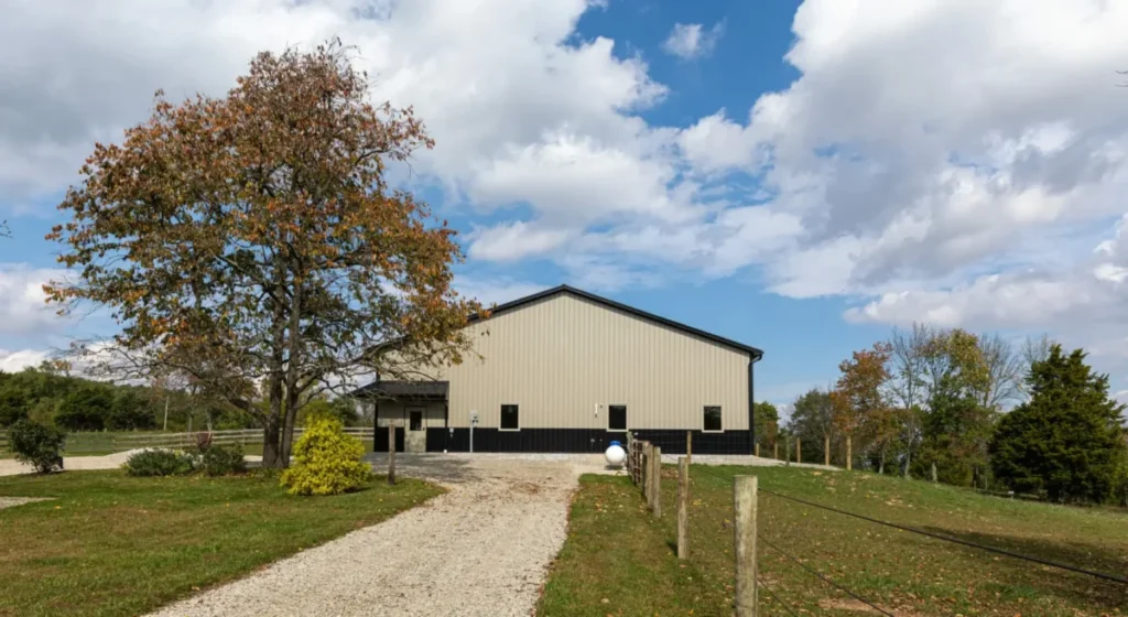 A large tan and black metal-sided pole barn building with a peaked roof sits at the end of a gravel driveway, surrounded by green grass, wooden fence posts, and autumn-colored trees under a partly cloudy sky in rural Bainbridge, Ohio.