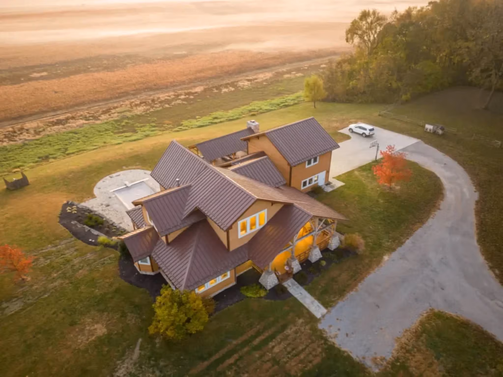Aerial drone view of a large multi-gabled home with dark brown standing seam metal roofing, warm amber exterior walls, a pool patio, and a curved driveway surrounded by autumn farmland and trees.