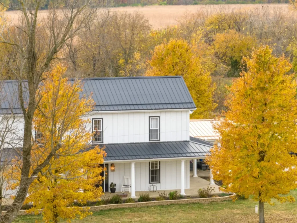 A two-story white board-and-batten farmhouse with charcoal standing seam metal roof and covered front porch, surrounded by golden yellow autumn trees with harvested fields in the background in rural Ohio.