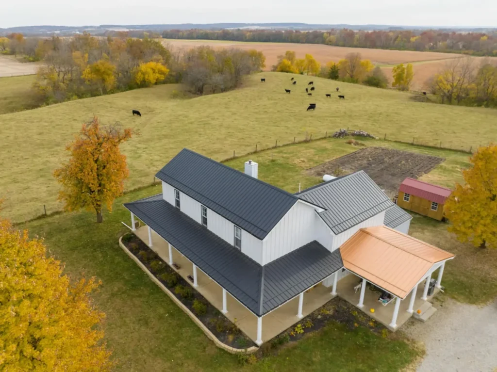 Aerial drone photograph of a modern white board-and-batten farmhouse featuring a dark charcoal standing seam metal roof and a copper-toned metal porch roof extension, surrounded by autumn-colored trees and rolling green pastures with black Angus cattle grazing in the background.