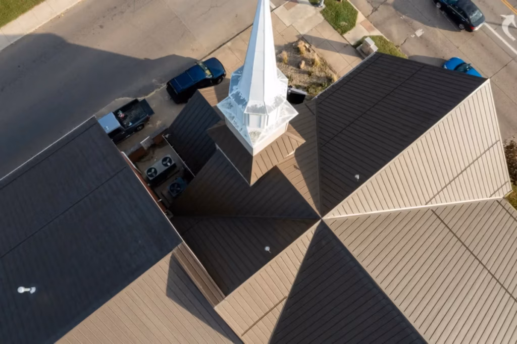 Aerial drone view of a church building featuring a tan and dark brown standing seam metal roof with a white painted steeple at the center, surrounded by a parking lot with black and blue vehicles.