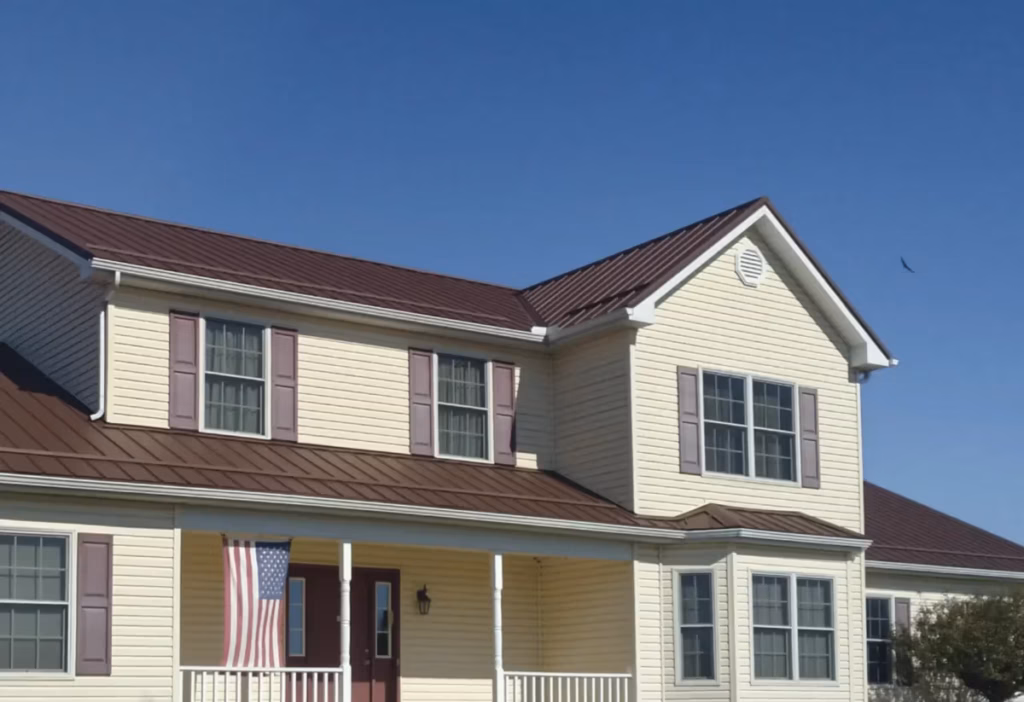 A two-story colonial-style house with cream vinyl siding, purple shutters, and a dark burgundy standing-seam metal roof, featuring a covered front porch with white columns and an American flag displayed near the front door.