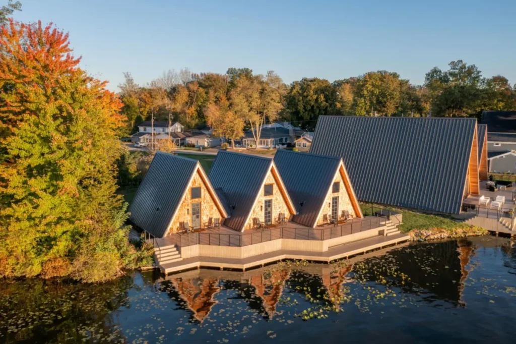 Aerial view of three A-frame cabins with dark metal standing-seam roofs and stone facades built on a waterfront deck over a calm lake, surrounded by autumn foliage in shades of orange, red, and gold.