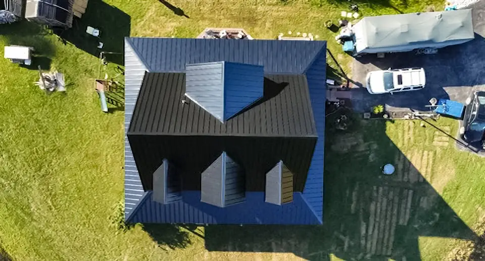 Aerial drone view of a residential home with a newly installed dark charcoal standing seam metal roof, surrounded by green grass yard with neighboring structures visible.