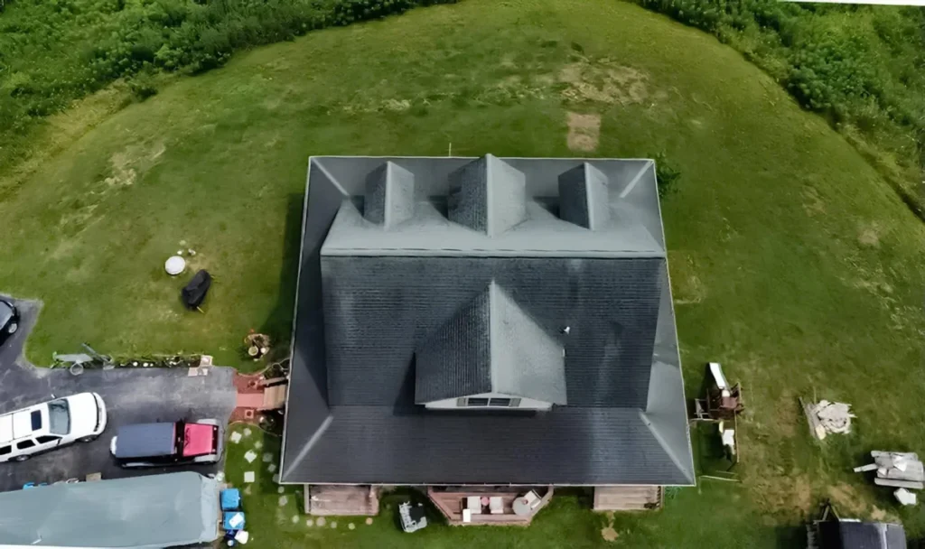 Aerial drone view of a large home with a newly installed dark charcoal gray metal roof surrounded by green grass, a paved driveway with parked vehicles, and a backyard deck.