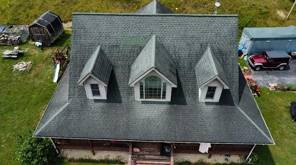 Aerial drone view of a large residential home with a dark gray asphalt shingle roof featuring three dormer windows, a covered front porch, and surrounding green yard in a rural setting.