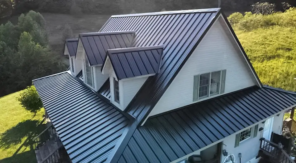 Aerial view of a white two-story farmhouse with a newly installed dark charcoal standing seam metal roof featuring multiple gabled dormers, surrounded by green hills and trees.