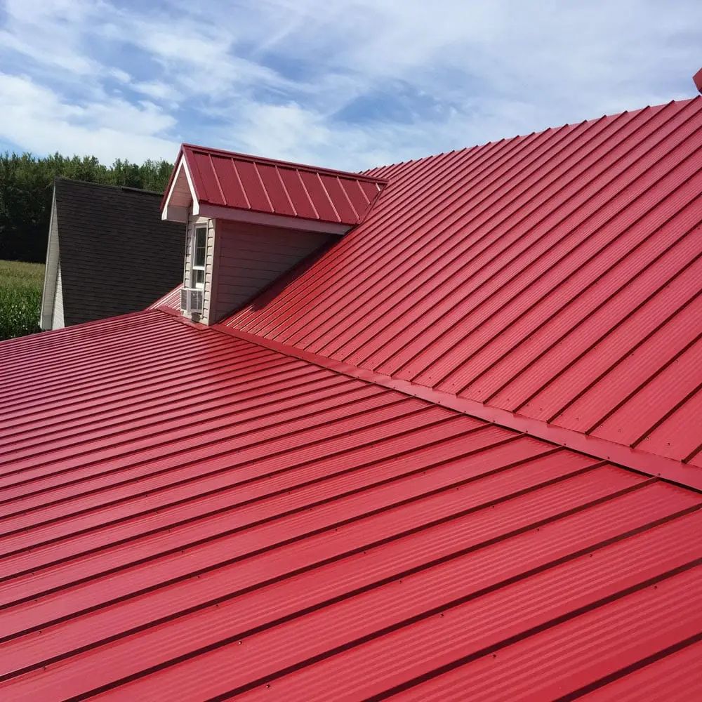 A bright red corrugated metal panel roof with multiple intersecting roof planes and a small white-trimmed dormer window, set against a partly cloudy sky with green trees in the background.