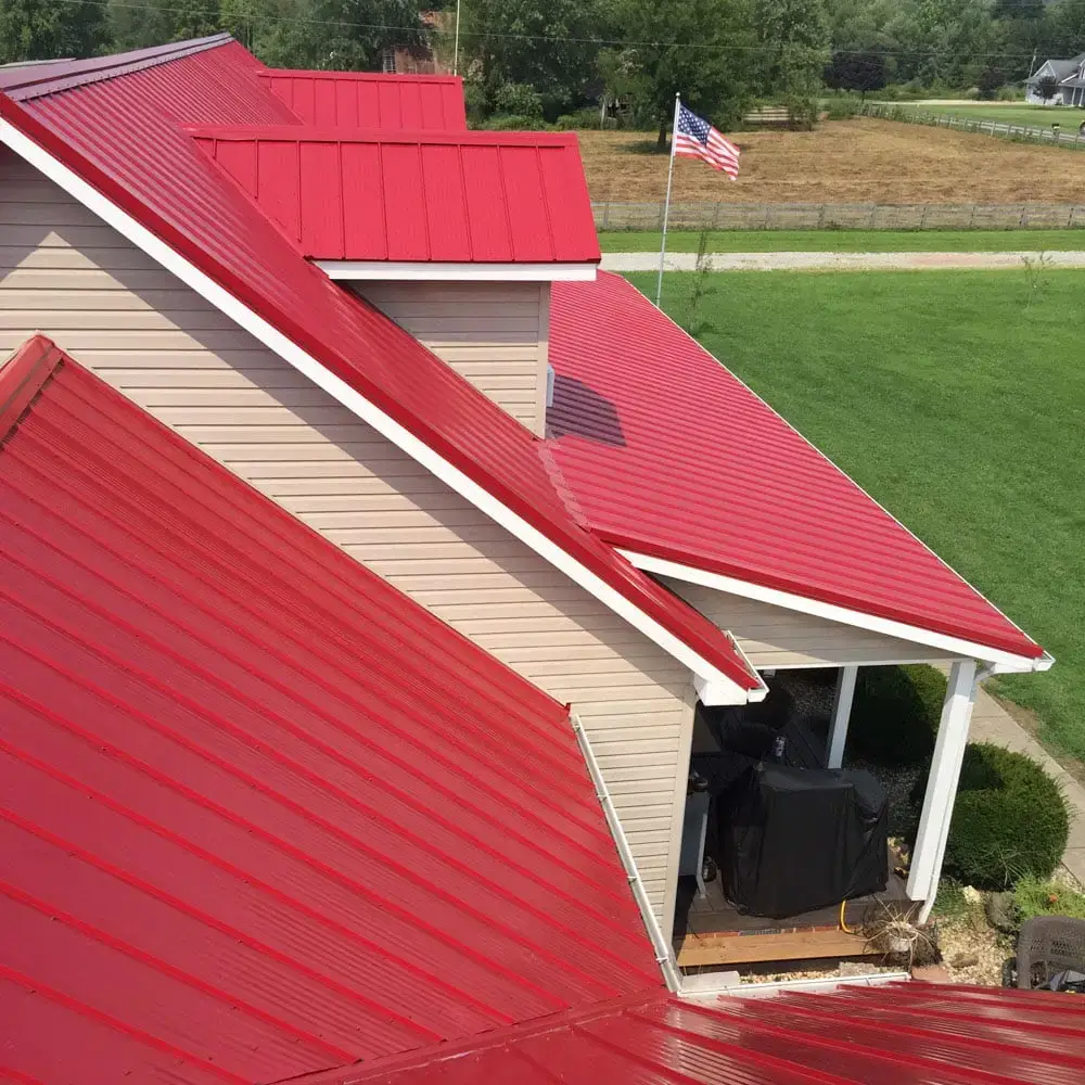 Aerial view of a residential home with a bold red standing seam metal roof featuring multiple roof lines, beige vinyl siding, a covered back porch with a black grill, and an American flag visible in the background against a green rural landscape.