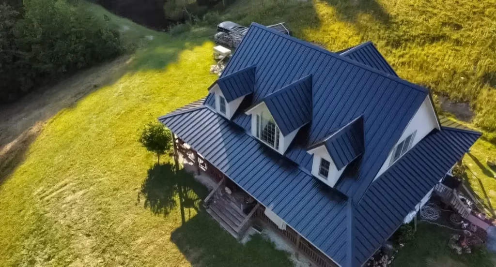Aerial drone view of a large country home with a dark navy blue standing seam metal roof, white dormers, and a wraparound porch, surrounded by lush green grass and trees.