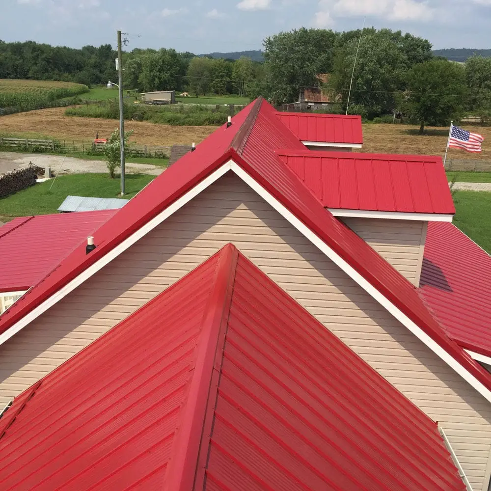 Aerial view of a large building with bright red standing-seam metal roofing panels and tan vinyl siding, surrounded by green farmland, cornfields, and an American flag flying in the background.