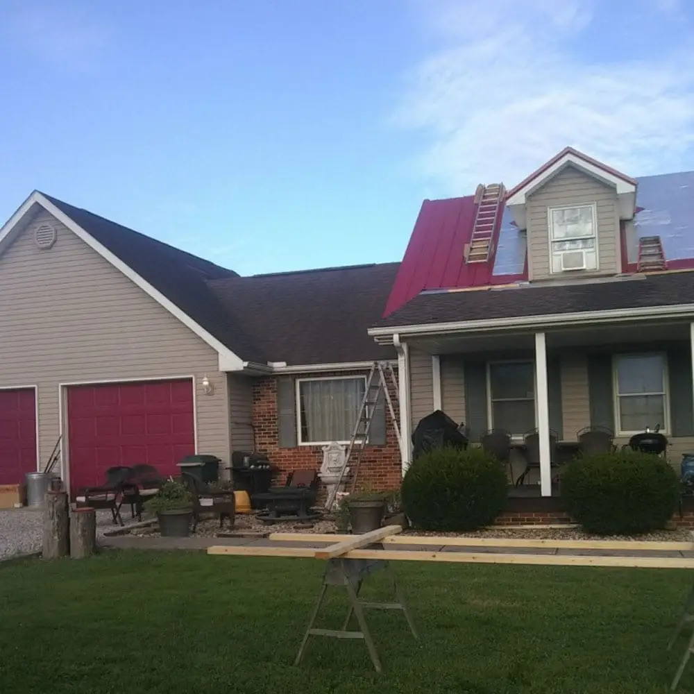 A residential ranch-style home with tan siding and brick facade undergoing a red metal roof installation, with ladders propped against the house, a sawhorse in the foreground, and red garage doors visible on the attached garage.
