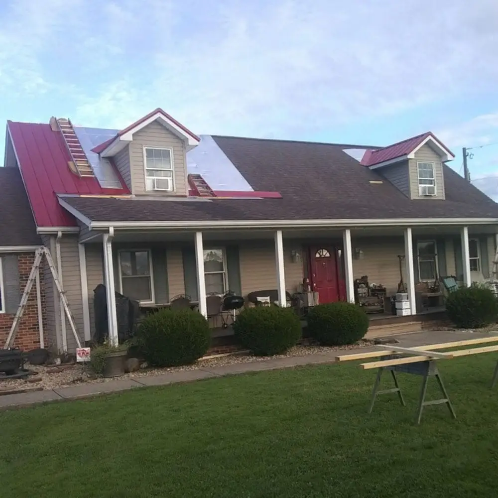 A one-story farmhouse with a covered front porch and red front door undergoing a red metal roof installation, with portions of the old dark shingle roof still visible alongside newly installed crimson metal panels and a ladder leaning against the left side.