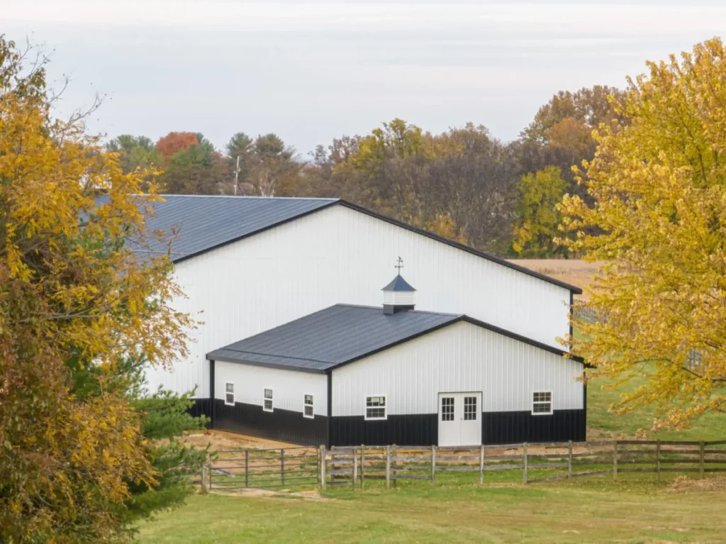 A large white metal pole barn with a dark charcoal standing-seam metal roof, black lower wainscoting, white-trimmed windows, French entry doors, and a decorative cupola with weather vane, surrounded by autumn-colored trees and split-rail fencing in a rural Ohio farm setting.