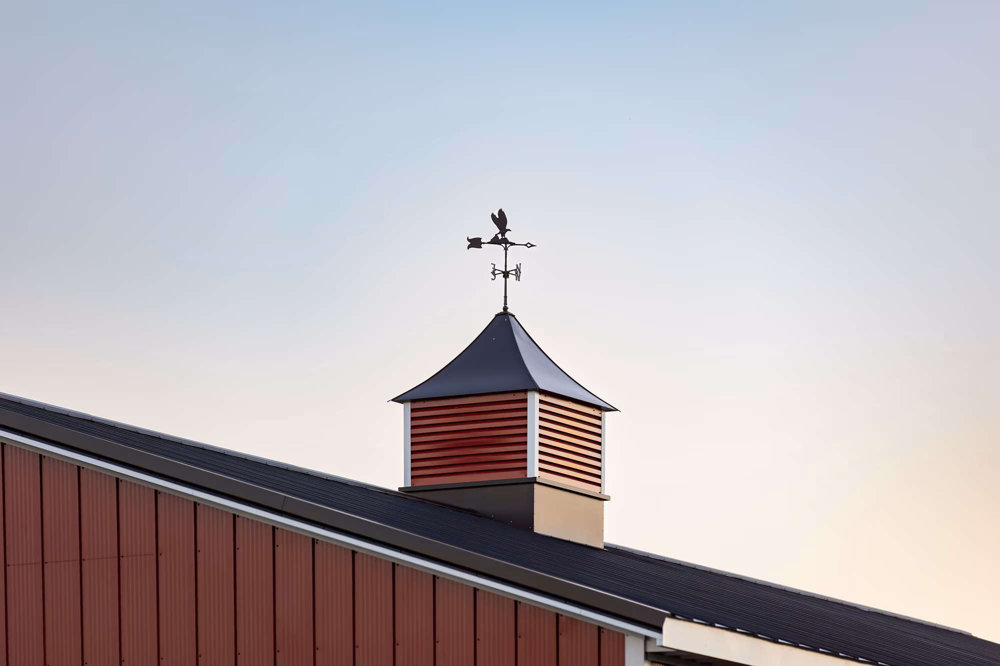 Red metal cupola with cross weathervane mounted on a dark metal roof edge against a light sky