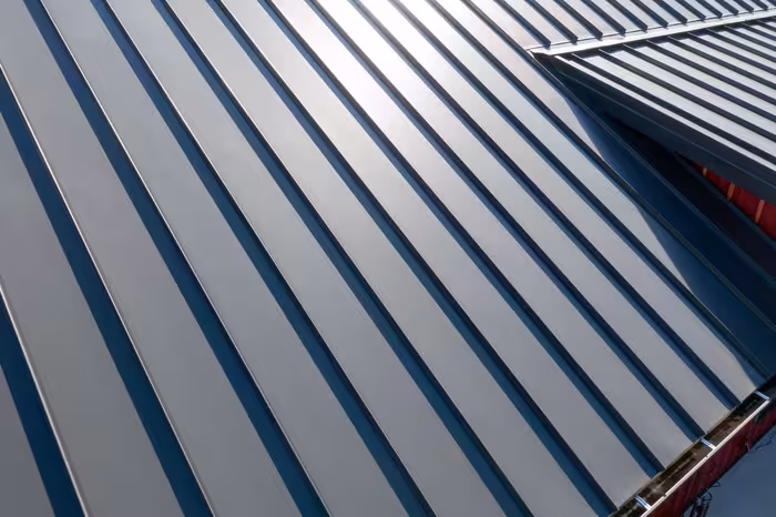 Close-up of metal roof panels with dark ribs and light gray finish, casting sharp shadows in sunlight.