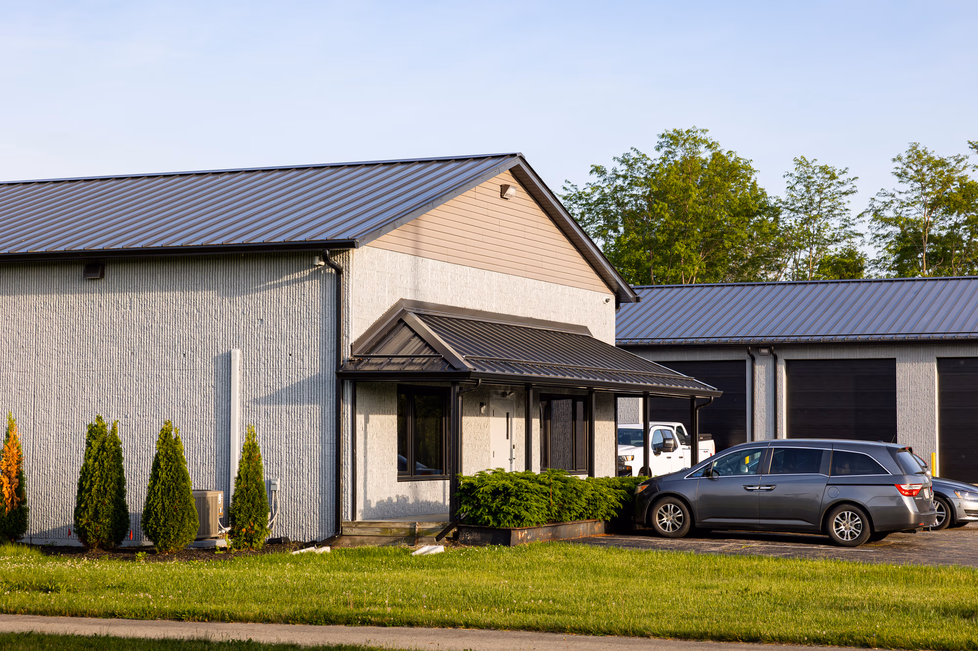 Modern commercial building featuring gray metal roofing and light-colored metal siding with dark trim, landscaped grounds, and parked vehicle.