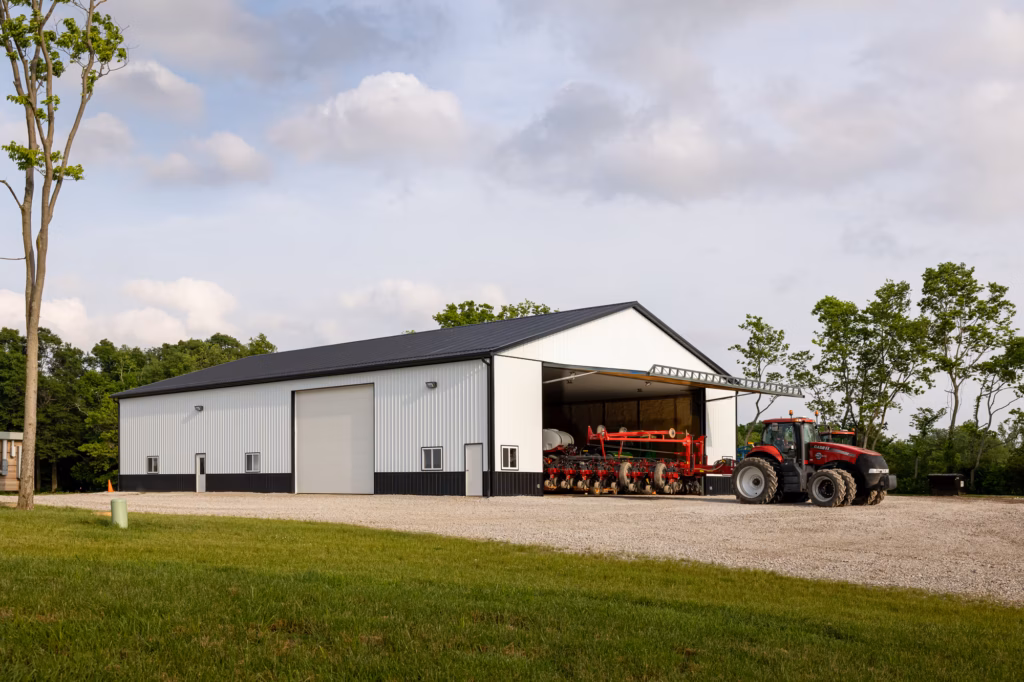 White metal-sided agricultural building with a red tractor and equipment parked outside on a paved area, surrounded by green grass and trees.