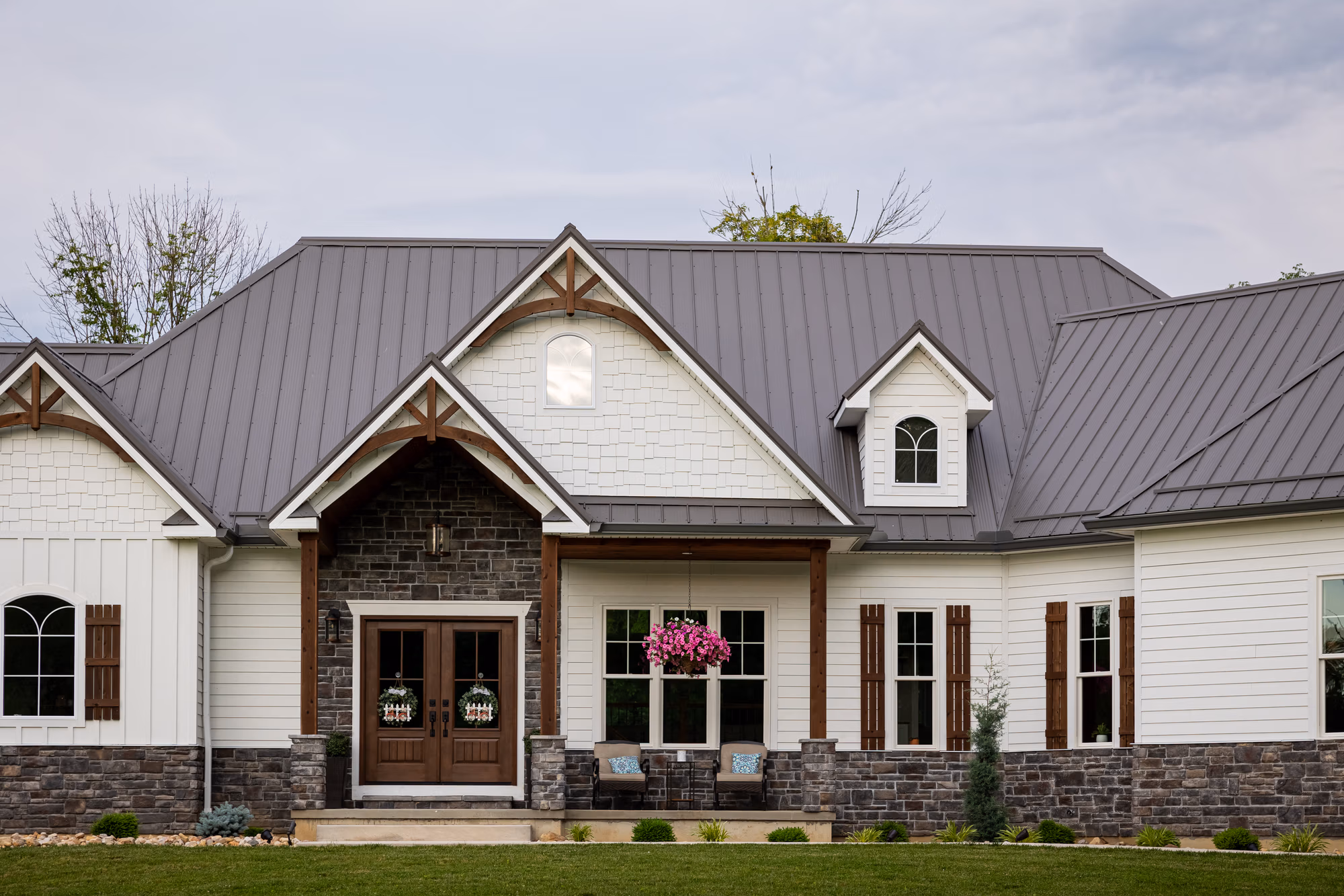 White house with stone base, metal roof with rippled Wave panel design, gray metal standing seam accents, and manicured landscaping