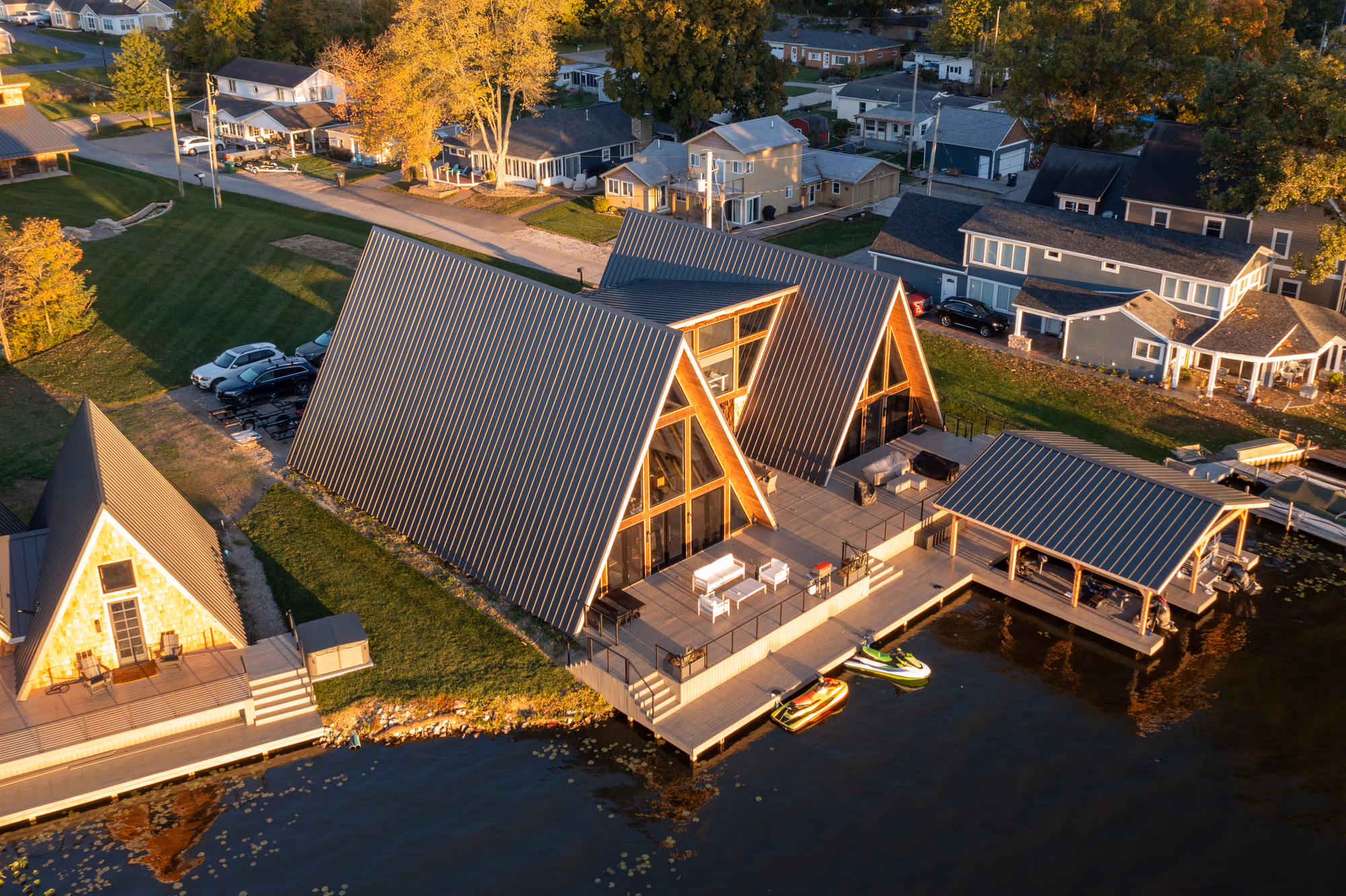 Modern A-frame home with dark standing seam metal roof alongside a pond, illuminated by warm evening light